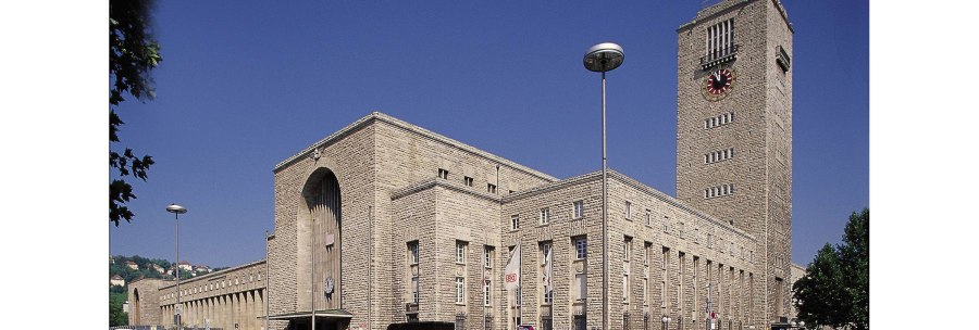 Der Hauptbahnhof Stuttgart mit seinem markanten Uhrturm und Mercedes-Stern, umgeben von Autos und Bäumen, unter einem klaren blauen Himmel., © Stuttgart-Marketing GmbH
