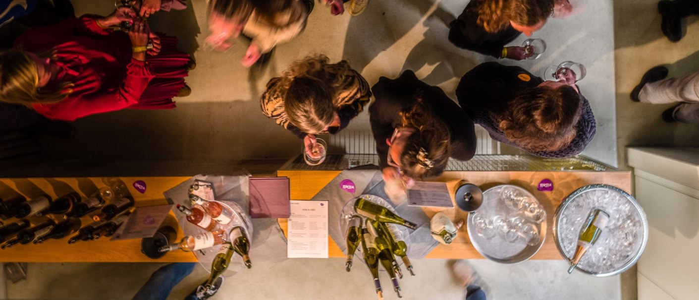 Menschen bei einer Weinverkostung, von oben fotografiert. Auf dem Tisch stehen Weinflaschen und Gläser. Die Atmosphäre wirkt gesellig und entspannt., © club traube