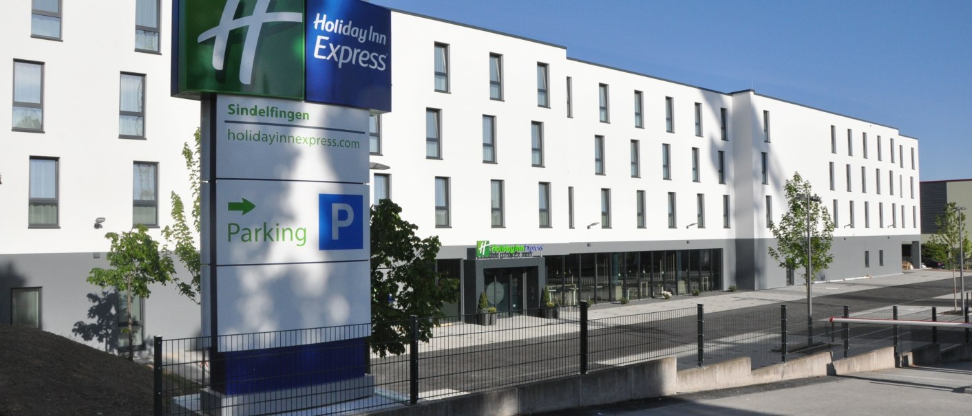The Holiday Inn Express in Sindelfingen has a modern façade with a large parking lot sign in the foreground. The sky is clear and blue., © tristar GmbH The Holiday Inn Express in Sindelfingen has a modern façade with a large parking lot sign in the foreground. The sky is clear and blue., © tristar GmbH