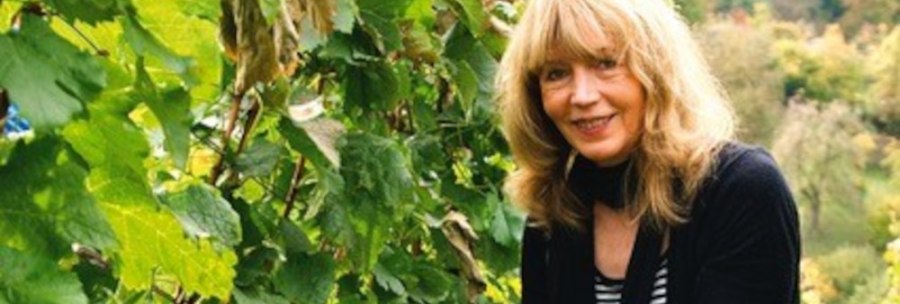 A woman with blonde hair harvests grapes in a green vineyard. She wears gloves and smiles at the camera., &copy; Elsbeth Koch