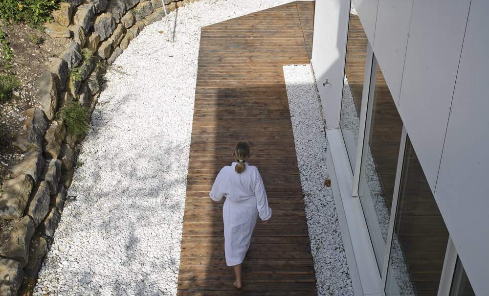 Eine Person im Bademantel geht auf einem Holzweg neben einem modernen Gebäude. Der Weg ist von weißen Steinen und einer Steinmauer umgeben., © Badhotel Restaurant Stauferland
