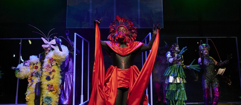 Personen in farbenfrohen, fantasievollen Kostümen und Masken auf einer Bühne, beleuchtet von buntem Licht., © Württembergische Staatstheater Stuttgart Personen in farbenfrohen, fantasievollen Kostümen und Masken auf einer Bühne, beleuchtet von buntem Licht., © Württembergische Staatstheater Stuttgart