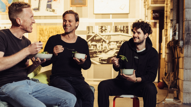 Three men sit together laughing and eating salad in a cozy, artistically designed room., © Vhy! GmbH Three men sit together laughing and eating salad in a cozy, artistically designed room., © Vhy! GmbH