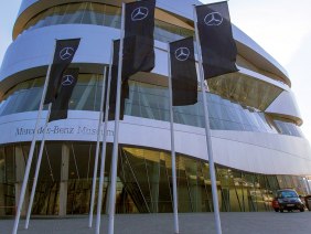 Das Mercedes-Benz Museum mit seiner modernen Architektur und mehreren Fahnen im Vordergrund. Der Himmel ist klar und blau., © SMG, Achim Mende