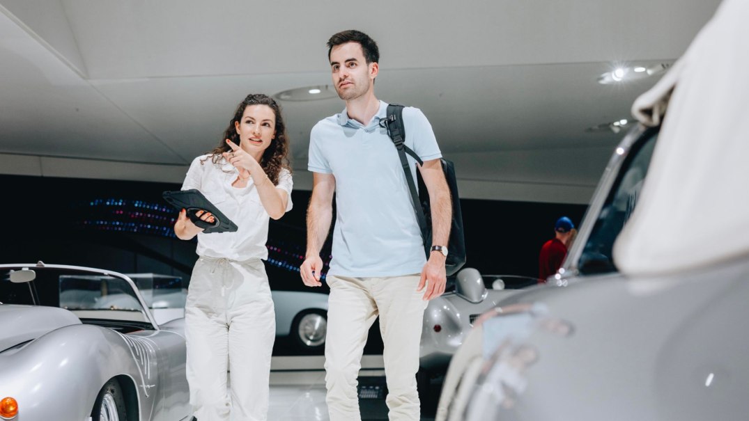 Zwei Personen in einem modernen Raum mit Autos. Eine Person zeigt auf etwas, während die andere ein Tablet hält. Im Hintergrund sind Autos zu sehen., © Porsche AG