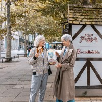 Zwei Frauen lachen vor einem Brezelstand am Schlossplatz. Eine hält eine Brezel, die andere eine Tüte. Herbstliche Bäume im Hintergrund., © SMG, Sarah Schmid