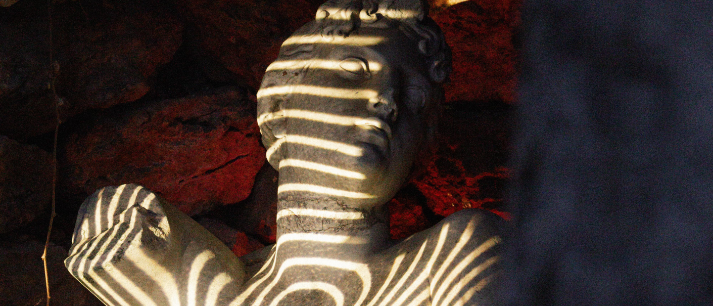 Eine Steinbüste mit Streifen aus Licht und Schatten, umgeben von einem rot-braunen Hintergrund., © Lichtgestalten