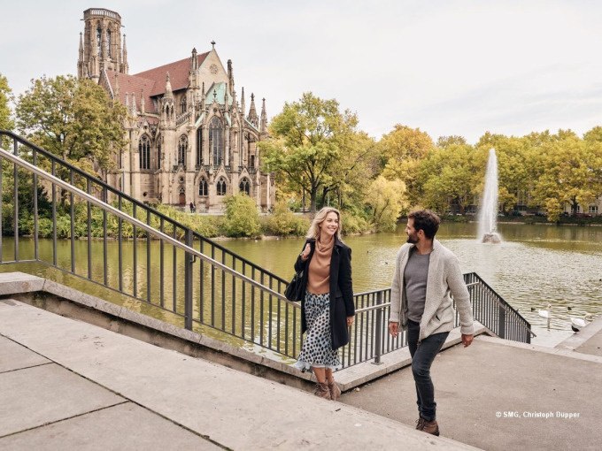 Zwei Personen gehen eine Treppe hinunter, im Hintergrund eine Kirche am Seeufer und ein Springbrunnen. Herbstliche B&auml;ume umgeben die Szene., &copy; Stuttgart Marketing GmbH