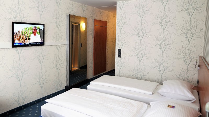 Hotelzimmer mit zwei Einzelbetten, einem Fernseher an der Wand und einem großen Spiegel. Die Wände sind mit einem baumartigen Muster tapeziert., © TOMAS