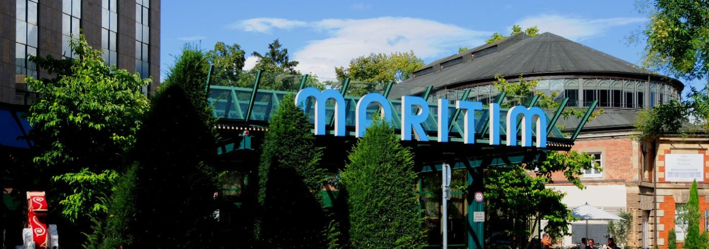 Au&szlig;enansicht des Maritim Hotels mit markantem Glasdach und umgeben von B&auml;umen und Pflanzen. Ein Weg f&uuml;hrt am Geb&auml;ude entlang., &copy; Maritim Hotelgesellschaft mbH
