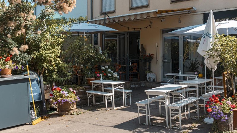 Gem&uuml;tlicher Au&szlig;enbereich eines Caf&eacute;s mit wei&szlig;en Tischen und St&uuml;hlen, umgeben von &uuml;ppigen Pflanzen und bunten Blumen., &copy; Stuttgart Marketing GmbH, Sarah Schmid