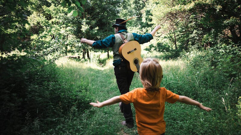 Ein Mann mit Hut und Gitarre auf dem Rücken führt ein Kind durch einen sonnigen Waldweg. Beide breiten die Arme aus., © FITZ Das Theater animierter Formen Ein Mann mit Hut und Gitarre auf dem Rücken führt ein Kind durch einen sonnigen Waldweg. Beide breiten die Arme aus., © FITZ Das Theater animierter Formen