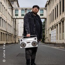 Eine Person steht in einer leeren Straße zwischen Gebäuden und hält eine Boombox. Die Szene wirkt urban und industriell., © links im Bild Eine Person steht in einer leeren Straße zwischen Gebäuden und hält eine Boombox. Die Szene wirkt urban und industriell., © links im Bild