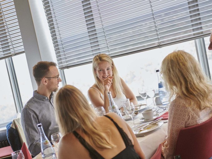 Vier Personen frühstücken an einem Tisch in einem Restaurant mit großen Fenstern. Sie unterhalten sich und genießen die Mahlzeit., © SWR Media Services GmbH / SWR Fernsehturm Stuttgart