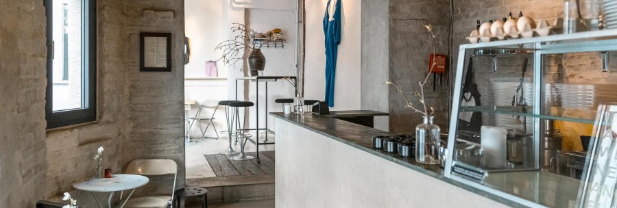Modern café with minimalist décor, white walls and wooden furniture. A blue piece of clothing hangs on the wall, plants on tables., © SMG Stuttgart Marketing GmbH - Sarah Schmid