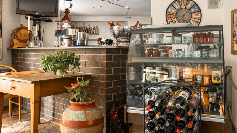 Gemütliches Café mit Holztisch, Weinregal, dekorativen Bildern und Pflanzen. Eine Theke mit Getränken und ein Fernseher sind ebenfalls zu sehen., © SMG, Sarah Schmid Gemütliches Café mit Holztisch, Weinregal, dekorativen Bildern und Pflanzen. Eine Theke mit Getränken und ein Fernseher sind ebenfalls zu sehen., © SMG, Sarah Schmid