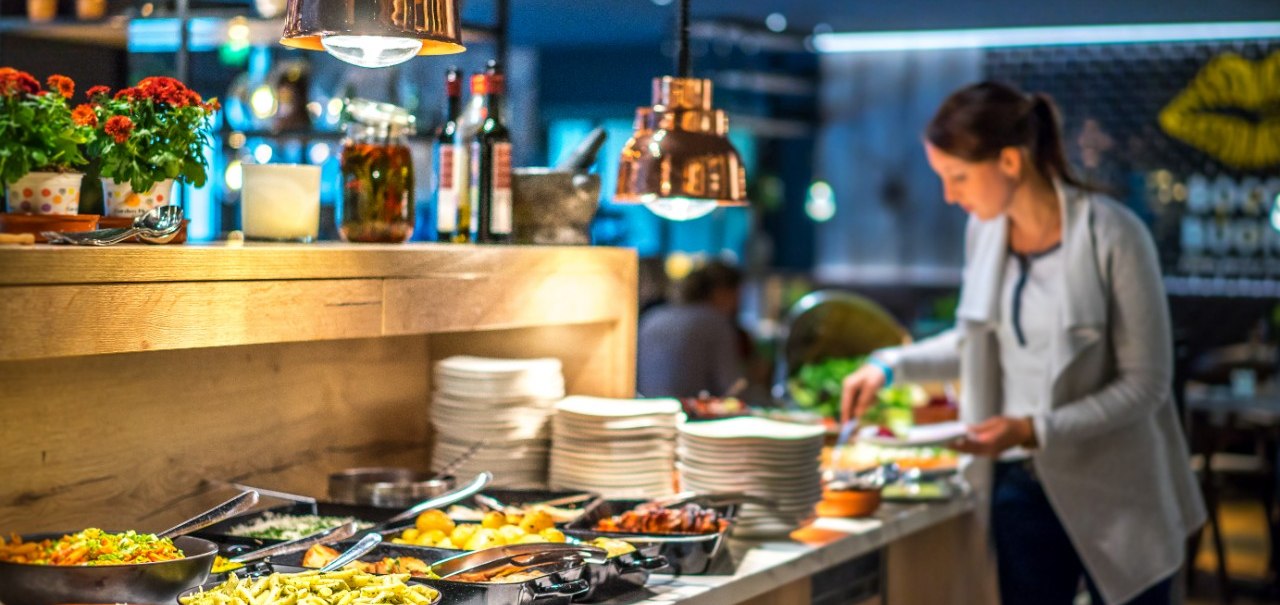 Eine Frau steht an einem Buffet in einem stilvoll beleuchteten Restaurant. Verschiedene Speisen und Teller sind auf dem Tresen arrangiert., © Park Inn by Radisson Stuttgart