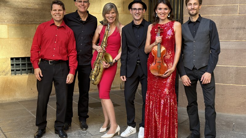 Sechs Personen in festlicher Kleidung posieren l&auml;chelnd. Zwei halten Musikinstrumente: ein Saxophon und eine Geige. Hintergrund aus Steinmauern., &copy; Friedrichsbau Variet&eacute;