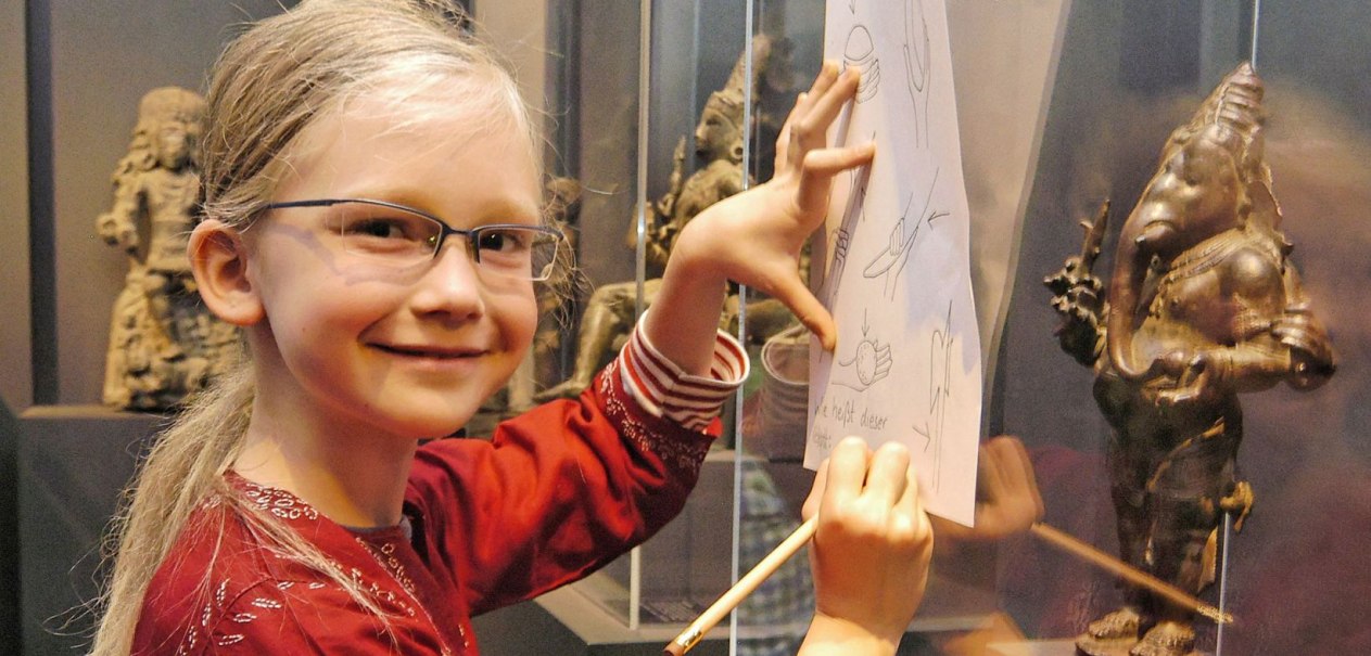 Ein Kind zeichnet in einem Museum neben einer s&uuml;dasiatischen Statue. Es tr&auml;gt eine Brille und ein rotes Oberteil., &copy; Linden-Museum Stuttgart