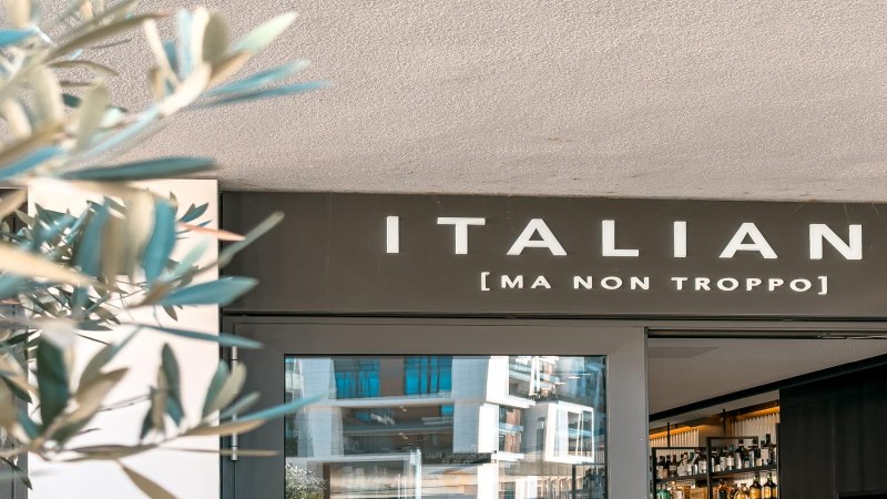 Entrance to a restaurant with the sign 'ITALIANI [MA NON TROPPO]'. Blurred plants can be seen in the foreground., &copy; SMG Stuttgart Marketing GmbH - Sarah Schmid