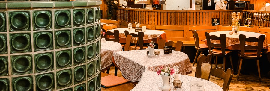 Cozy restaurant with wooden furniture, tablecloths and a green tiled stove. Lamps hanging from the ceiling, bar in the background., © Weinstube Kachelofen