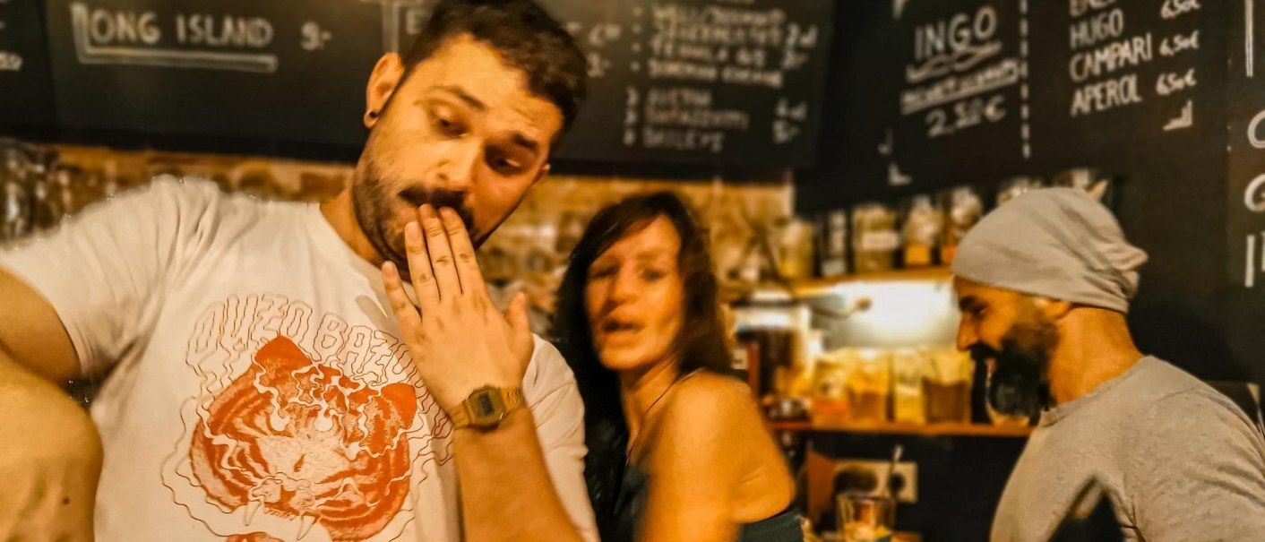 Drei Personen in einer Bar, eine mit Hand vor dem Mund, eine lächelnd, eine mit Mütze. Im Hintergrund eine Getränketafel., © Café Galao