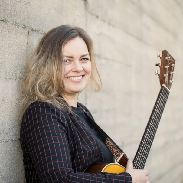 A smiling person is leaning against a wall and holding a guitar. They are wearing a plaid jacket., &copy; Renitenztheater Stuttgart e.V.