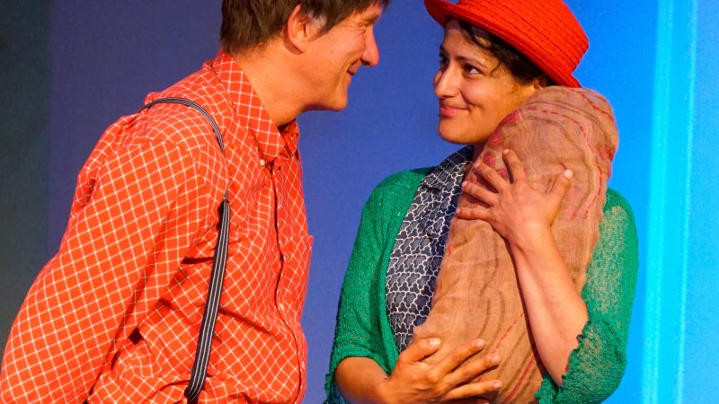 Zwei Personen in bunten Kost&uuml;men auf einer B&uuml;hne. Eine Person h&auml;lt ein B&uuml;ndel, beide l&auml;cheln sich an. Die Szene wirkt fr&ouml;hlich und lebendig., &copy; Katja Ritter