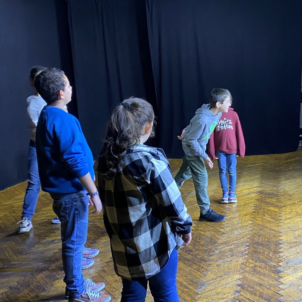 F&uuml;nf Kinder stehen auf einer B&uuml;hne mit dunklem Hintergrund, offenbar bei einer Theaterprobe. Sie tragen l&auml;ssige Kleidung und schauen in verschiedene Richtungen., &copy; Tathea