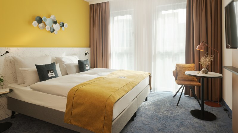 Modernes Hotelzimmer mit gelber Wand, Doppelbett, dekorativen Kissen, kleinem Tisch, Stuhl und Wand-TV. Helle Vorhänge lassen Licht herein., © the niu Kettle - Novum Hospitality Modernes Hotelzimmer mit gelber Wand, Doppelbett, dekorativen Kissen, kleinem Tisch, Stuhl und Wand-TV. Helle Vorhänge lassen Licht herein., © the niu Kettle - Novum Hospitality