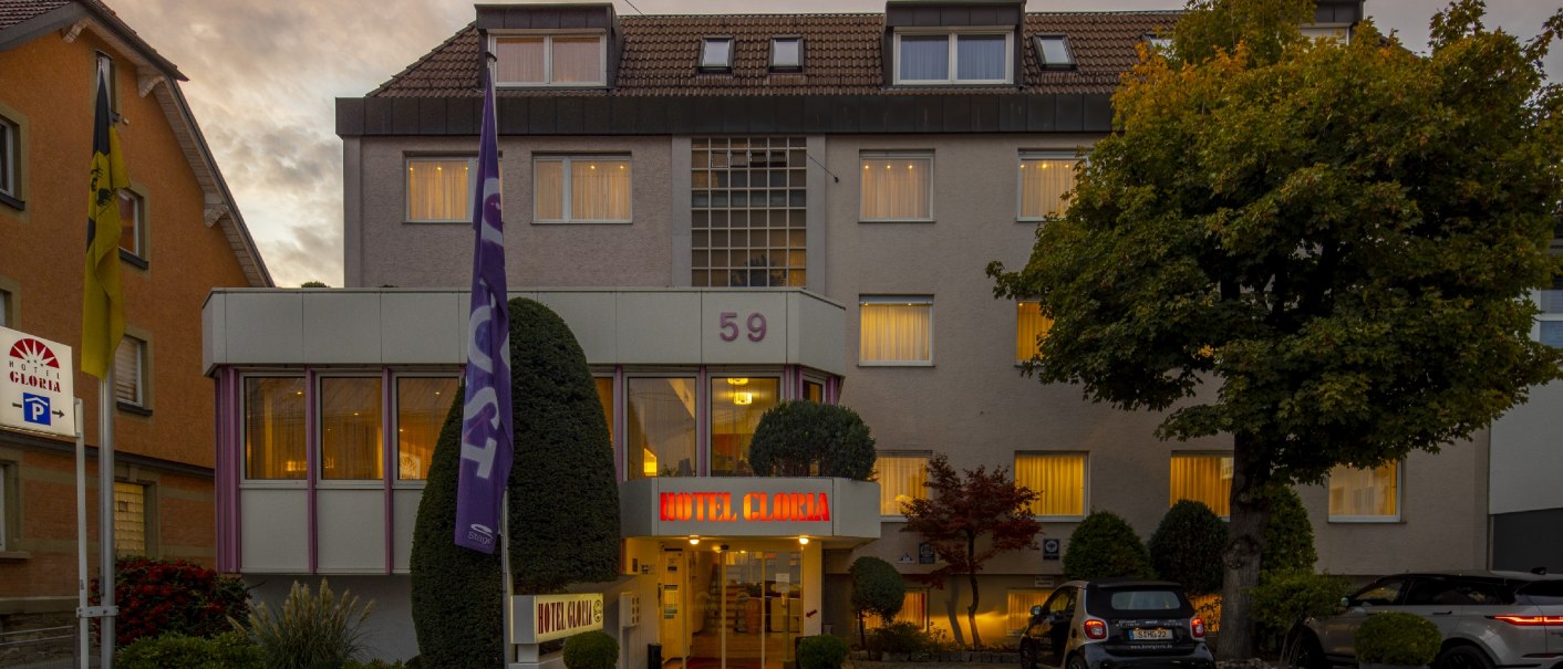Hotel building with illuminated windows and entrance, surrounded by trees and parked cars at dusk., © TOMAS Hotel building with illuminated windows and entrance, surrounded by trees and parked cars at dusk., © TOMAS