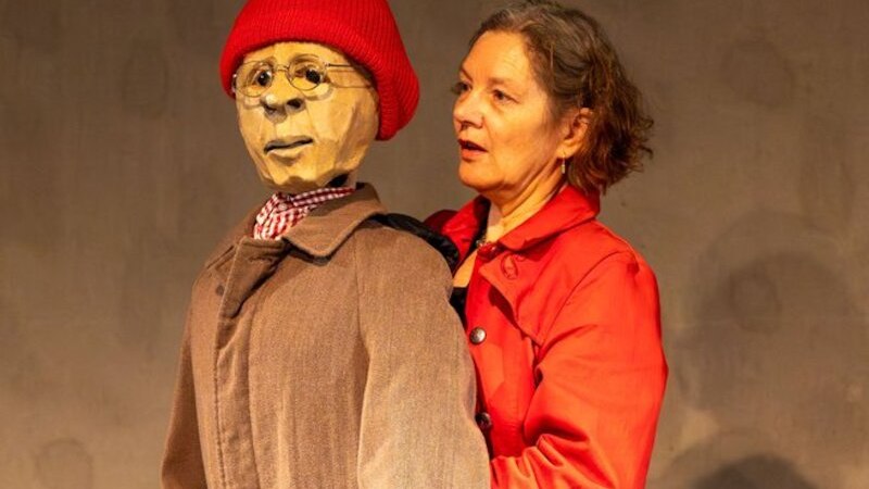 Eine Frau in roter Jacke interagiert mit einer Puppe, die eine rote M&uuml;tze und eine Brille tr&auml;gt. Die Szene wirkt theatralisch., &copy; Theaterhaus Stuttgart e.V.