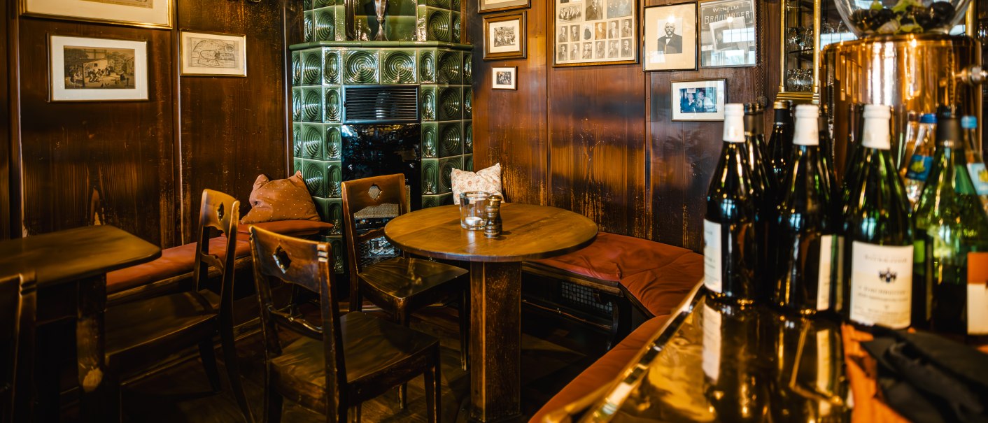 Gemütliche Weinstube mit Holzmöbeln, grünem Kachelofen, Bildern an den Wänden und Weinflaschen auf einem Tisch., © SMG Stuttgart Marketing GmbH - Sarah Schmid Gemütliche Weinstube mit Holzmöbeln, grünem Kachelofen, Bildern an den Wänden und Weinflaschen auf einem Tisch., © SMG Stuttgart Marketing GmbH - Sarah Schmid