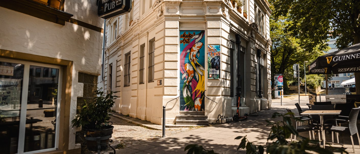 Eine Straßenecke mit einem Pub-Schild, bunter Wandkunst und Café-Tischen unter einem Sonnenschirm., © SMG Stuttgart Marketing GmbH - Sarah Schmid Eine Straßenecke mit einem Pub-Schild, bunter Wandkunst und Café-Tischen unter einem Sonnenschirm., © SMG Stuttgart Marketing GmbH - Sarah Schmid