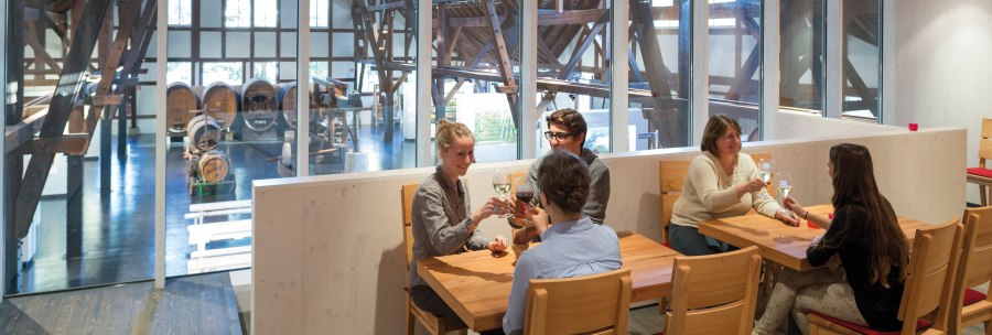 Menschen sitzen in einer modernen Vinothek im Weinbaumuseum Stuttgart, trinken Wein und unterhalten sich. Im Hintergrund sind große Weinfässer sichtbar., © Stuttgart-Marketing GmbH (SMG)