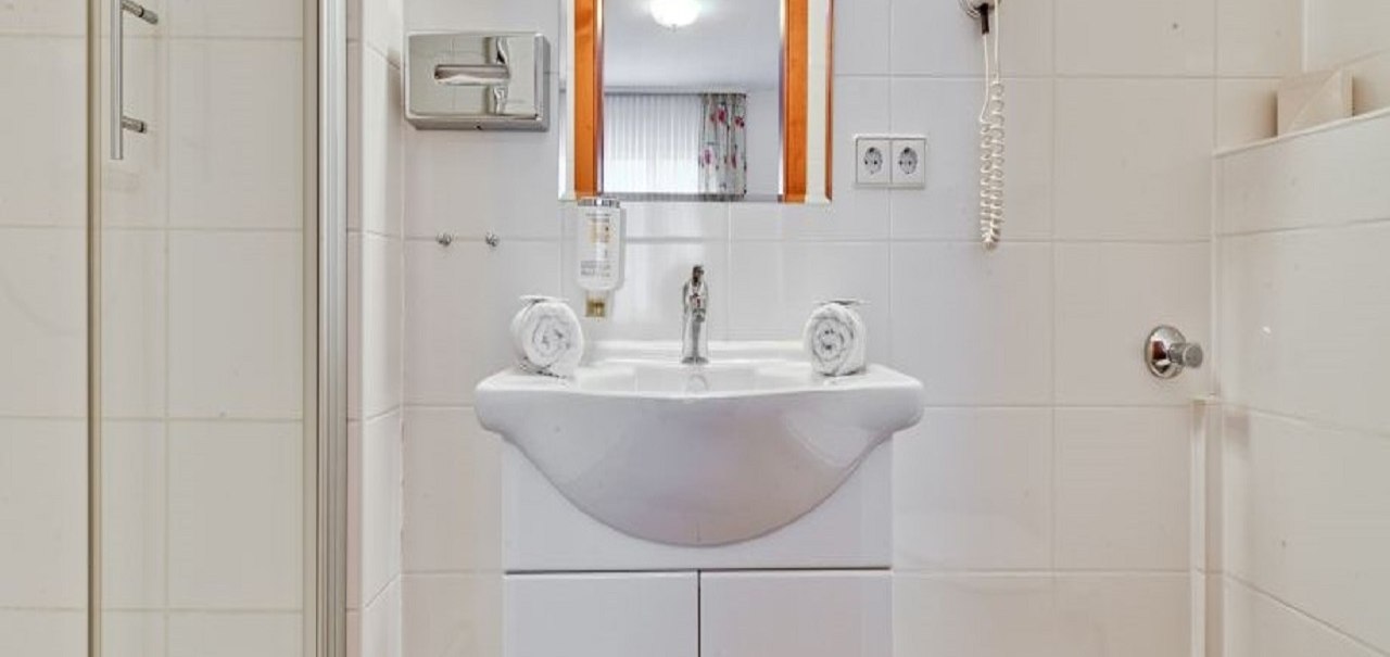 Ein modernes Badezimmer mit weißem Waschbecken, Spiegel, Haartrockner und Dusche. Handtücher sind ordentlich aufgerollt., © ARNOTEL GmbH