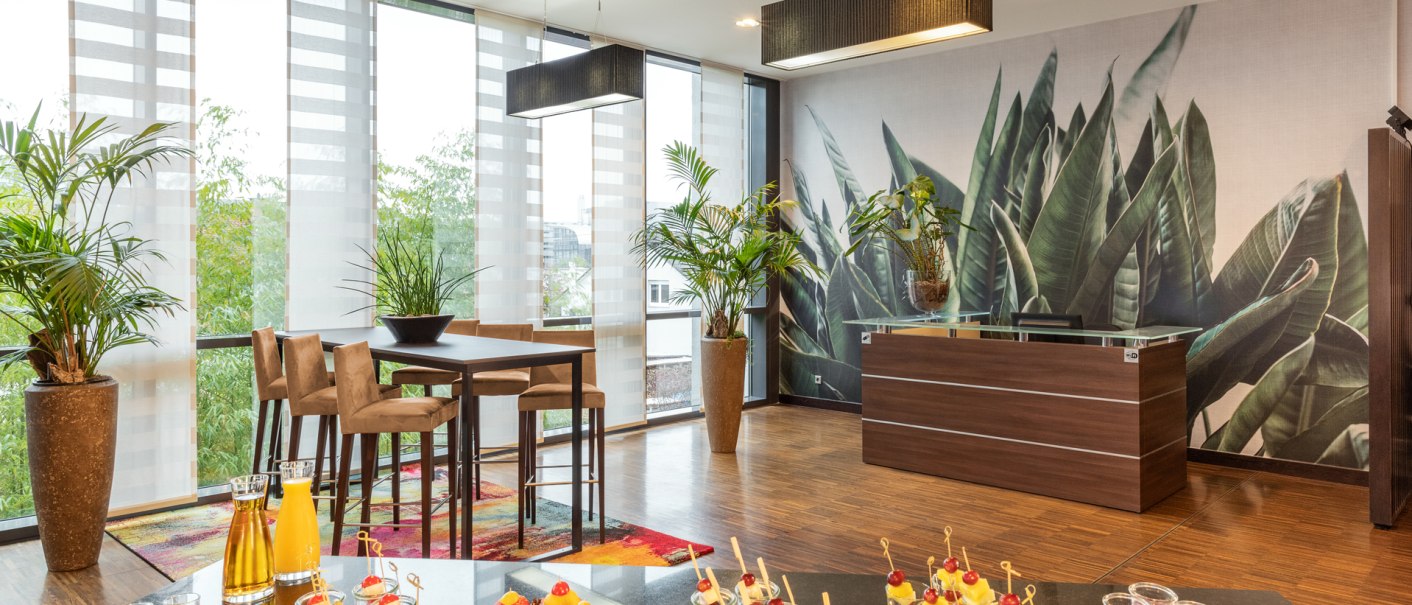 Moderner Kaffeepausenbereich mit hohen Tischen, Pflanzen und einem Buffet mit Snacks und Getränken. Große Fenster und Pflanzenmuster an der Wand., © NH Hotel Stuttgart Airport Moderner Kaffeepausenbereich mit hohen Tischen, Pflanzen und einem Buffet mit Snacks und Getränken. Große Fenster und Pflanzenmuster an der Wand., © NH Hotel Stuttgart Airport