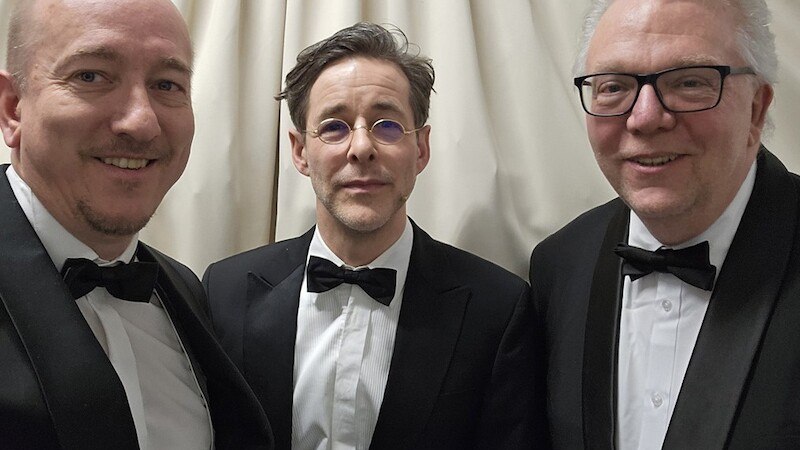 Drei Männer in eleganten Anzügen und Fliegen posieren vor einem cremefarbenen Vorhang. Sie lächeln in die Kamera., © Theaterhaus Stuttgart e.V. Drei Männer in eleganten Anzügen und Fliegen posieren vor einem cremefarbenen Vorhang. Sie lächeln in die Kamera., © Theaterhaus Stuttgart e.V.