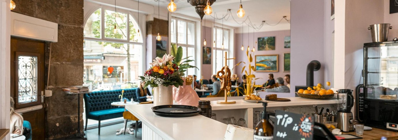 Gem&uuml;tliches Caf&eacute; mit Blumen und Brezeln auf der Theke, blauen Sofas und Kunst an den W&auml;nden. Gro&szlig;e Fenster lassen viel Licht herein., &copy; SMG, Sarah Schmid