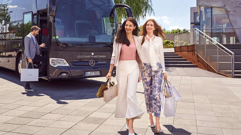 Zwei Frauen mit Einkaufstaschen gehen lächelnd von einem Bus weg. Ein Mann steigt in den Bus ein. Der Bus hat die Aufschrift 'Stuttgart ↔ Metzingen'., © Outletcity Metzingen GmbH