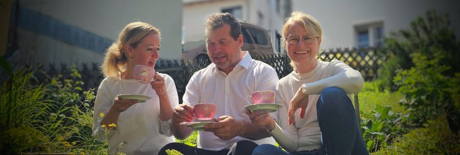 From left to right: Annette Ehrlich, Holger Köckritz and Annette Currle sit in the garden and enjoy tea from pink cups. They are wearing white tops and smiling at the camera., © Perle Uhlbach From left to right: Annette Ehrlich, Holger Köckritz and Annette Currle sit in the garden and enjoy tea from pink cups. They are wearing white tops and smiling at the camera., © Perle Uhlbach