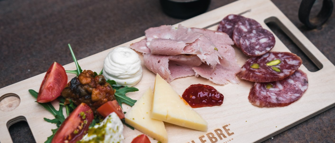 A wooden platter with ham, salami, cheese, tomatoes and rocket. Next to it is a bottle of beer labeled 'Bellabionda Helles'., © CELEBRE