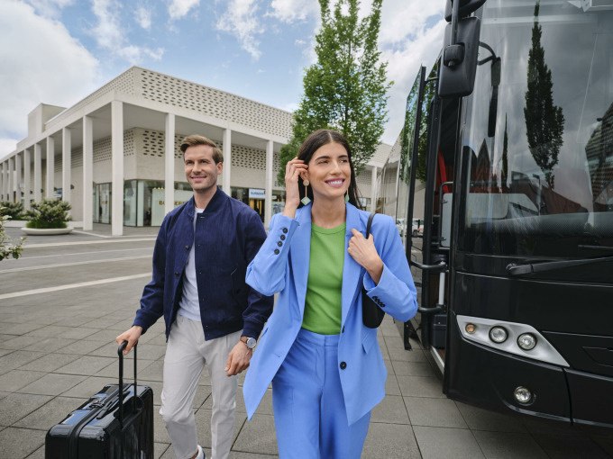 Zwei Personen steigen aus einem Bus an einer modernen Haltestelle aus. Sie tragen stilvolle Kleidung und haben Gepäck dabei., © Schlienz-Tours GmbH & Co.KG Zwei Personen steigen aus einem Bus an einer modernen Haltestelle aus. Sie tragen stilvolle Kleidung und haben Gepäck dabei., © Schlienz-Tours GmbH & Co.KG