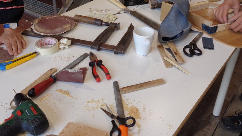 Werkbank mit verschiedenen Werkzeugen wie Bohrer, Zangen, Scheren und Holzstücken in einer Holzwerkstatt. Zwei Personen arbeiten an Projekten., © KVWH Werkbank mit verschiedenen Werkzeugen wie Bohrer, Zangen, Scheren und Holzstücken in einer Holzwerkstatt. Zwei Personen arbeiten an Projekten., © KVWH