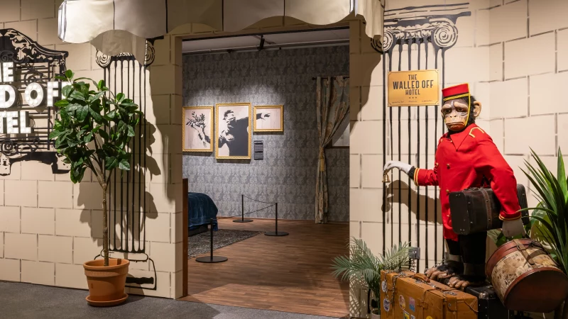 Eingang der Ausstellung „The Walled Off Hotel“ mit Banksy-Bezug, beleuchtetem Schild, Türöffnung, Koffer und Affenfigur in roter Uniform., © Dominik Gruss Eingang der Ausstellung „The Walled Off Hotel“ mit Banksy-Bezug, beleuchtetem Schild, Türöffnung, Koffer und Affenfigur in roter Uniform., © Dominik Gruss