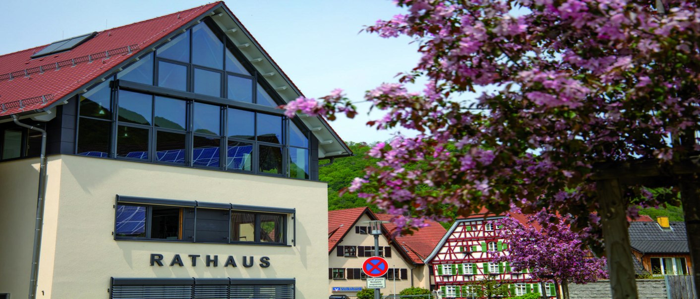 The modern town hall of Bad Ueberkingen with large windows and red roof. Flowering trees in the foreground, traditional half-timbered houses in the background., © © Stuttgart-Marketing GmbH