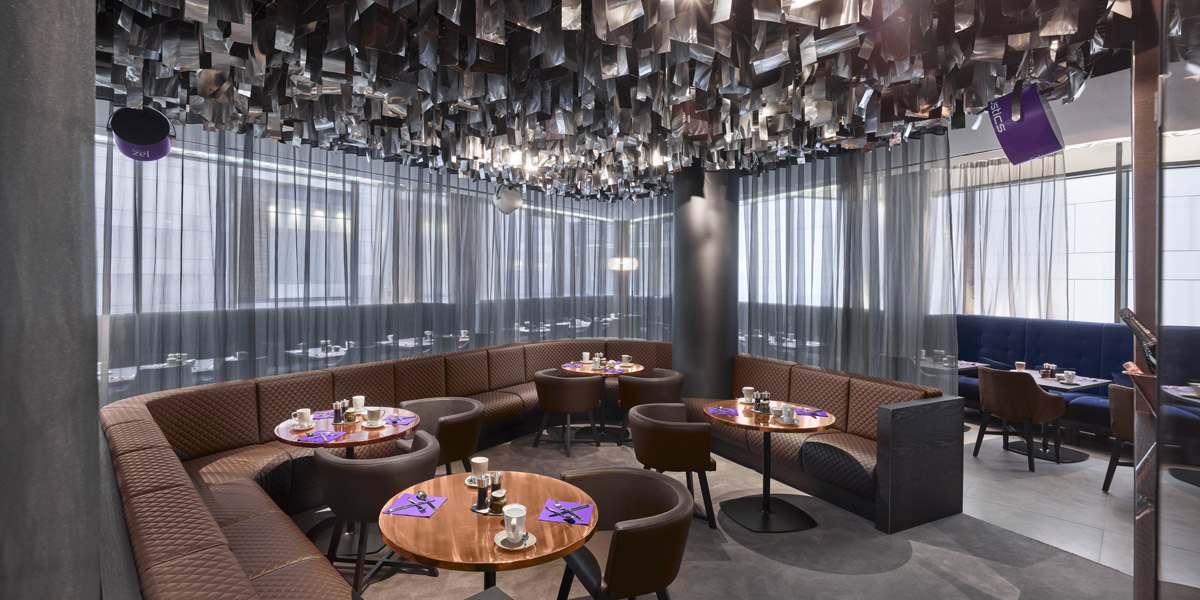 Modern restaurant with round wooden tables, brown chairs and decorative ceiling. Subdued lighting and curtains create a cozy atmosphere., © Jaz Hotel Stuttgart Modern restaurant with round wooden tables, brown chairs and decorative ceiling. Subdued lighting and curtains create a cozy atmosphere., © Jaz Hotel Stuttgart