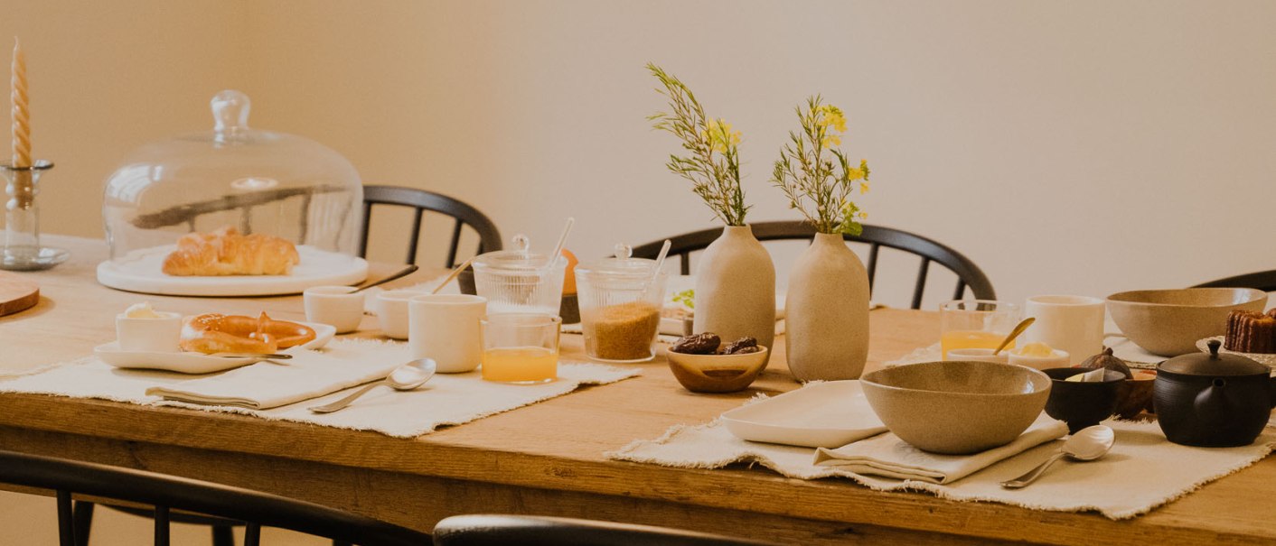 Ein Fr&uuml;hst&uuml;ckstisch mit Croissants, Saft, Geschirr und Blumenvasen. Eine H&auml;ngelampe beleuchtet den Tisch. Schwarze St&uuml;hle umgeben den Holztisch., &copy; Peter Oliver Wolff / DAS EDITH