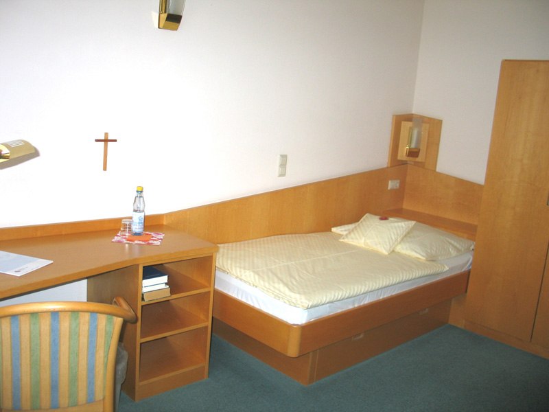 Ein schlichtes Einzelzimmer mit Holzmöbeln, Bett, Schreibtisch, Stuhl und einem Kreuz an der Wand., © Ev. Diakonissenanstalt Stuttgart Ein schlichtes Einzelzimmer mit Holzmöbeln, Bett, Schreibtisch, Stuhl und einem Kreuz an der Wand., © Ev. Diakonissenanstalt Stuttgart