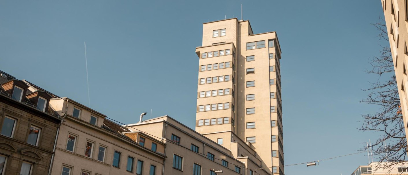 Der Tagblatt-Turm in Stuttgart ragt in den blauen Himmel, umgeben von älteren Gebäuden. Ein Kondensstreifen zieht sich durch den Himmel., © Stuttgart Marketing GmbH, Sarah Schmid